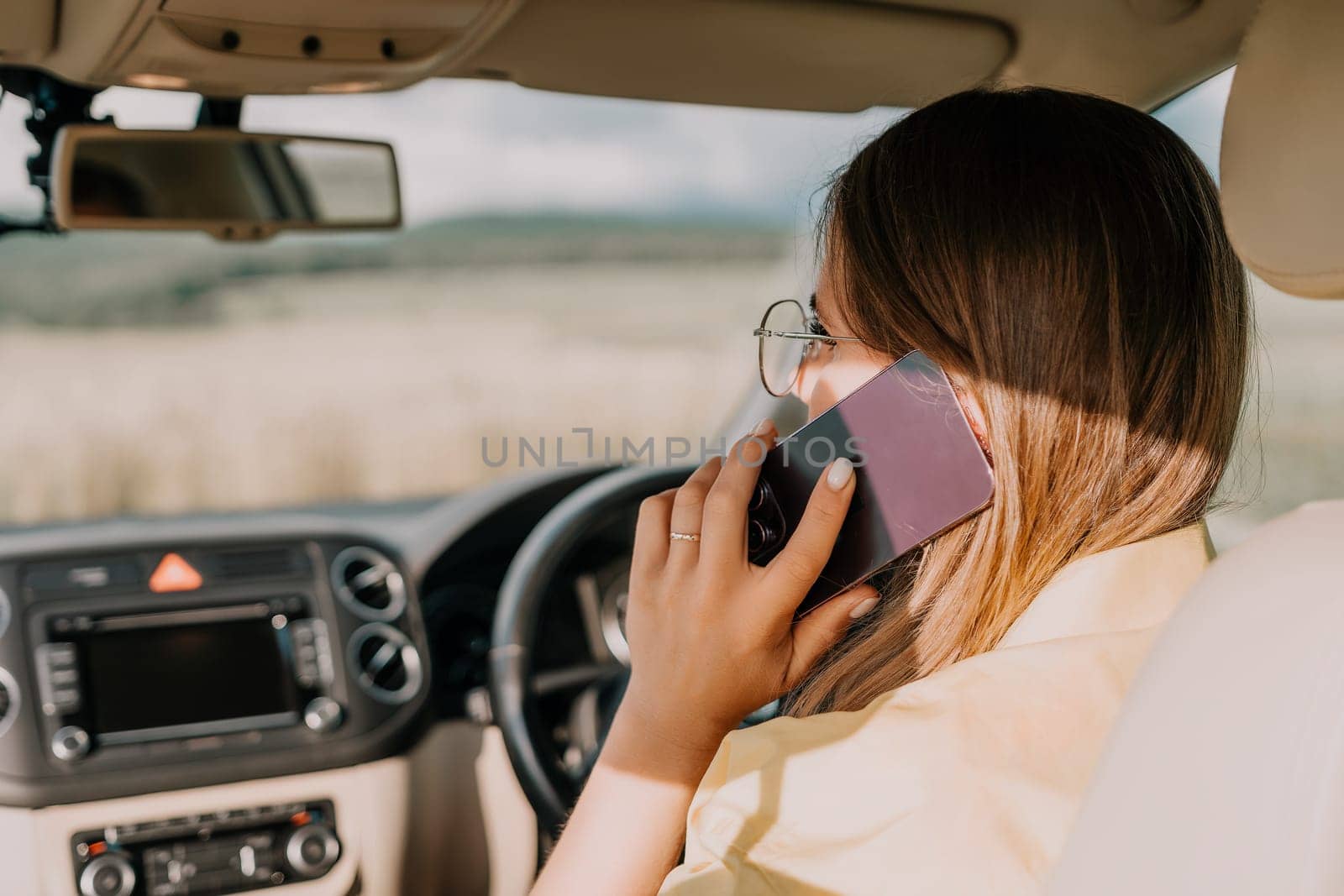 A woman in a yellow shirt is talking on the phone while driving a car. The car is in motion and the view outside the window is blurred.