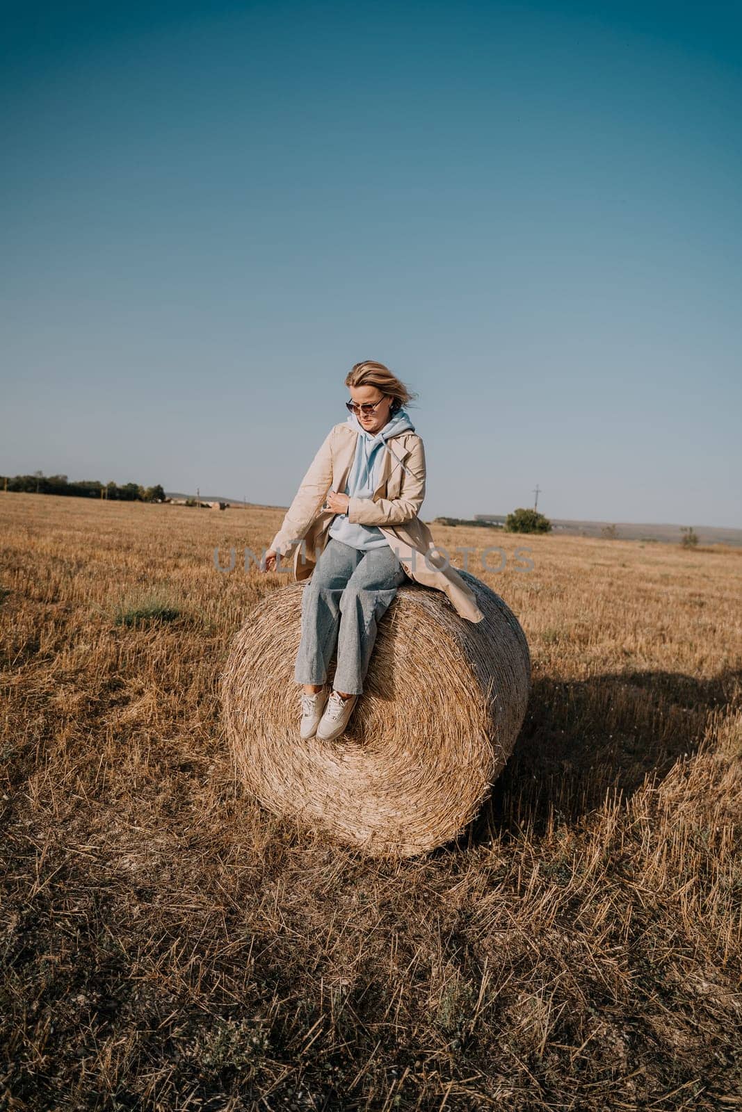 A woman in a beige coat and blue jeans sits on a hay bale in a field. The sky is a clear blue, and the field is brown and yellow.