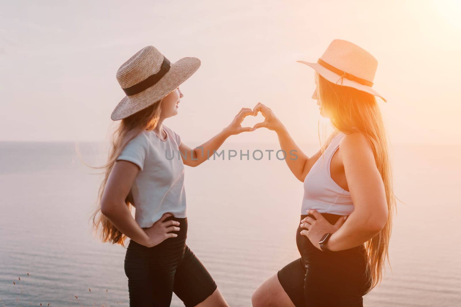 A mother and daughter stand facing each other on a cliff overlooking a body of water. They are wearing casual clothing and both have their hands up forming a heart shape.