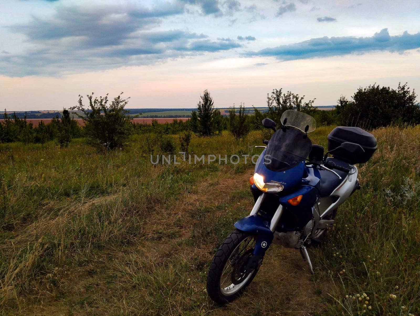 A motorcycle rests on grassy terrain as the sun sets, casting warm hues over the landscape with fields in the distance.