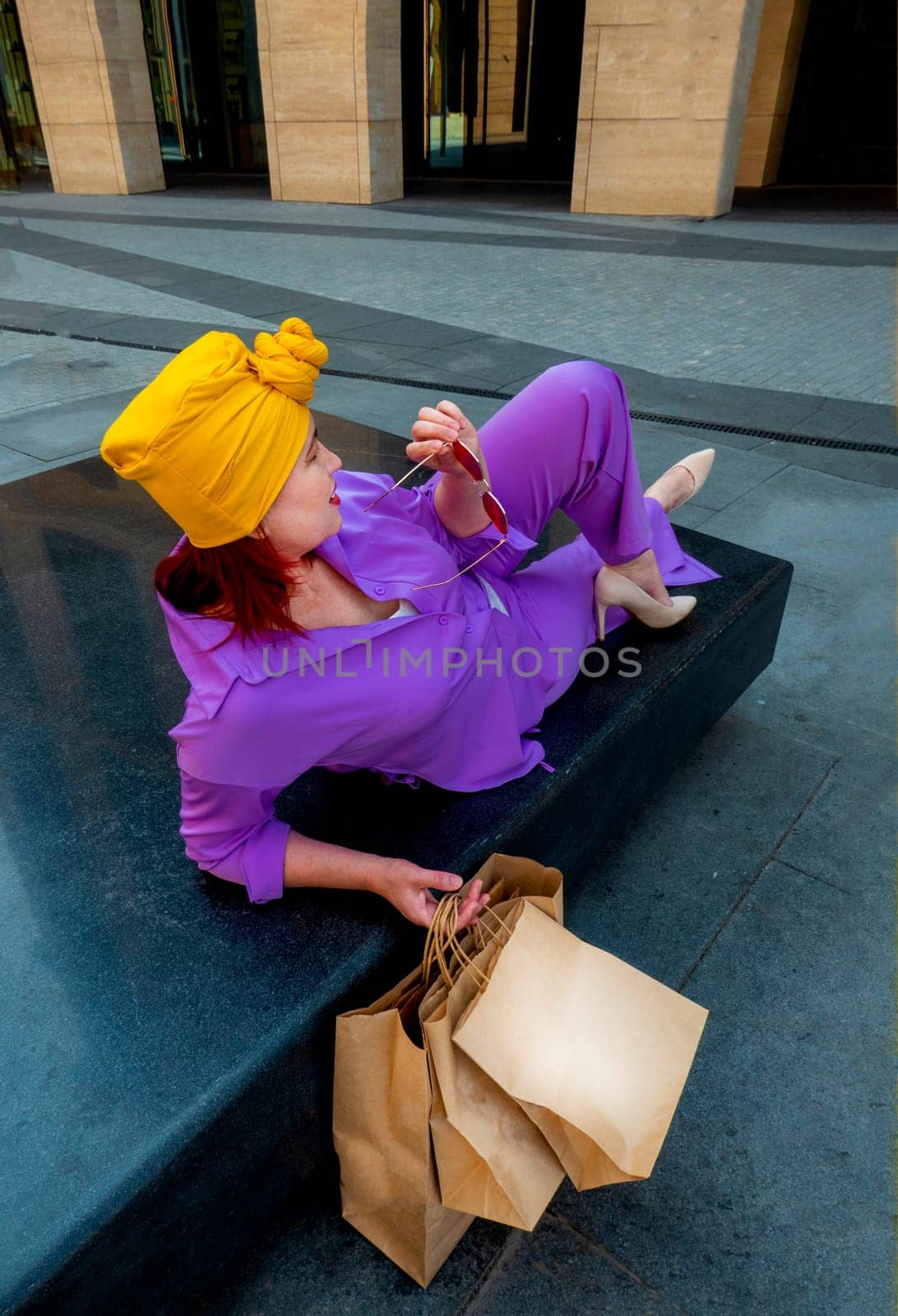woman with craft shopping bags in the city. The concept of fashionable Consumerism and ecology. Adult Asian woman in a purple suit with red and yellow accents enjoys shopping. rest between purchases
