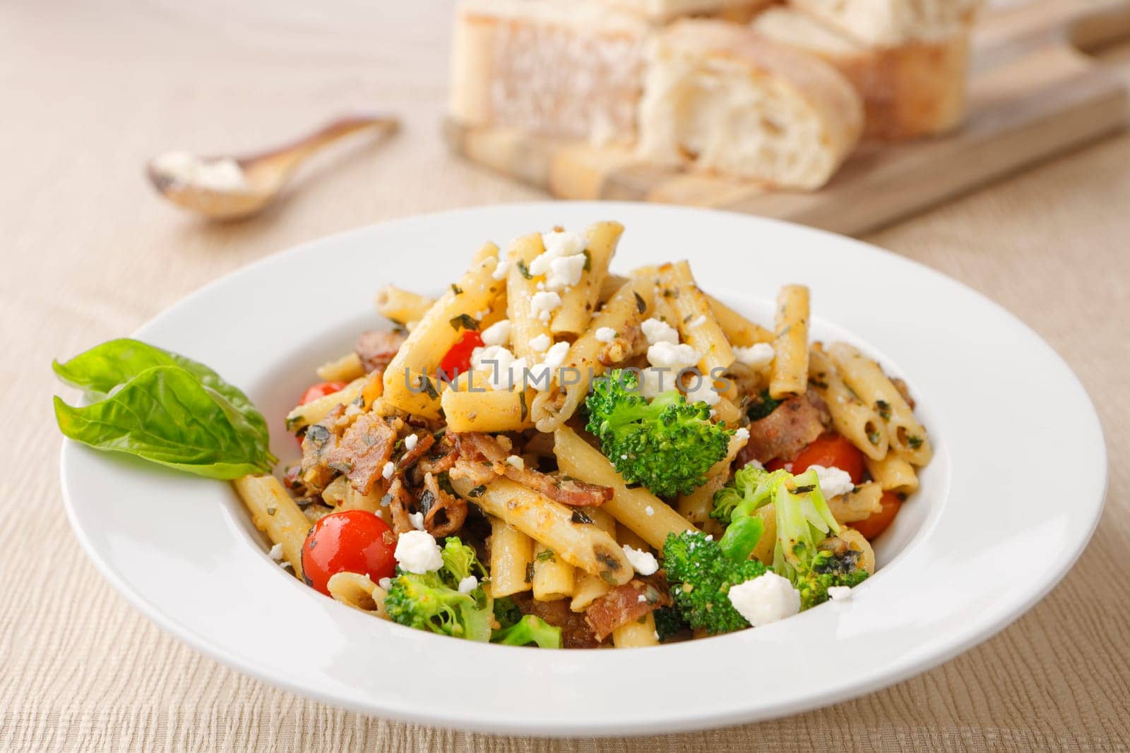 Studio photo of a bowl of penne pasta with broccoli and feta cheese and a cutting board of ciabatta bread slices.