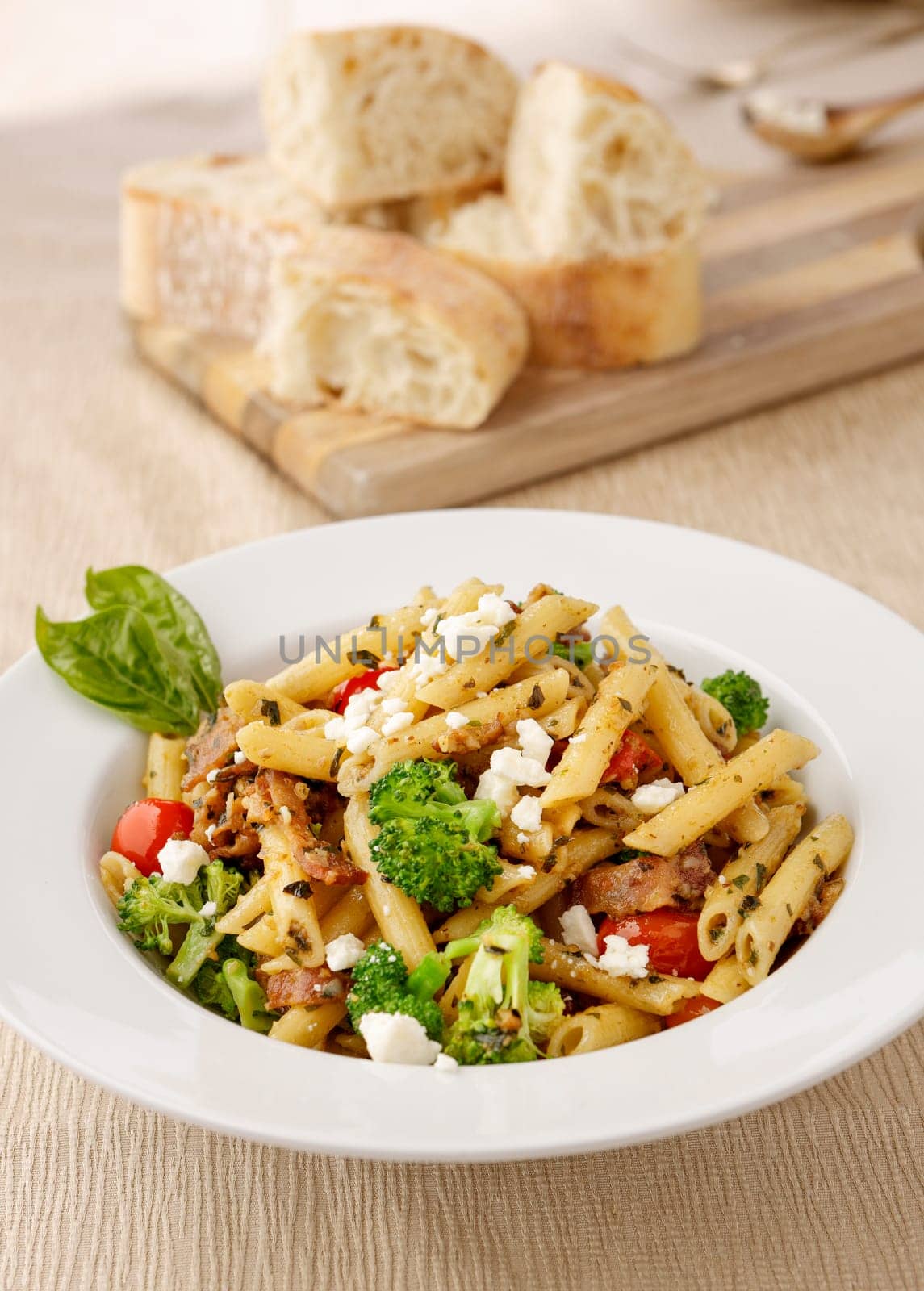 Studio photo of a bowl of penne pasta with broccoli and feta cheese and a cutting board of ciabatta bread slices.