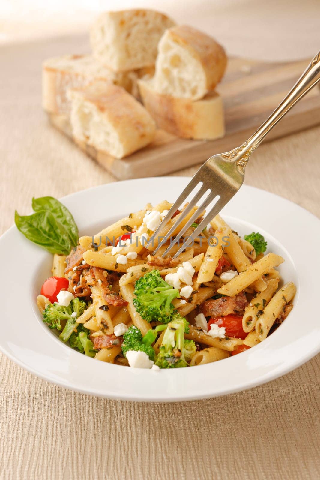 Studio photo of a fork going into a bowl of penne pasta with broccoli and feta cheese and a cutting board of ciabatta bread slices.