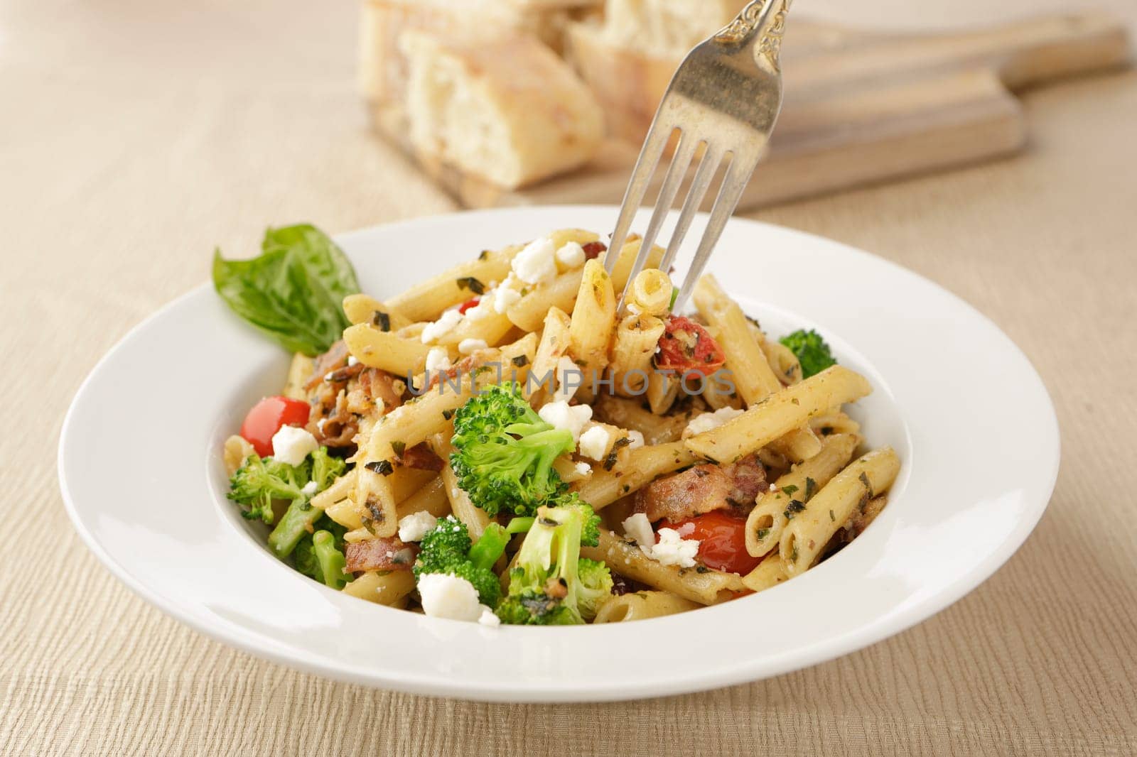 Studio photo of a fork going into a bowl of penne pasta with broccoli and feta cheese and a cutting board of ciabatta bread slices.