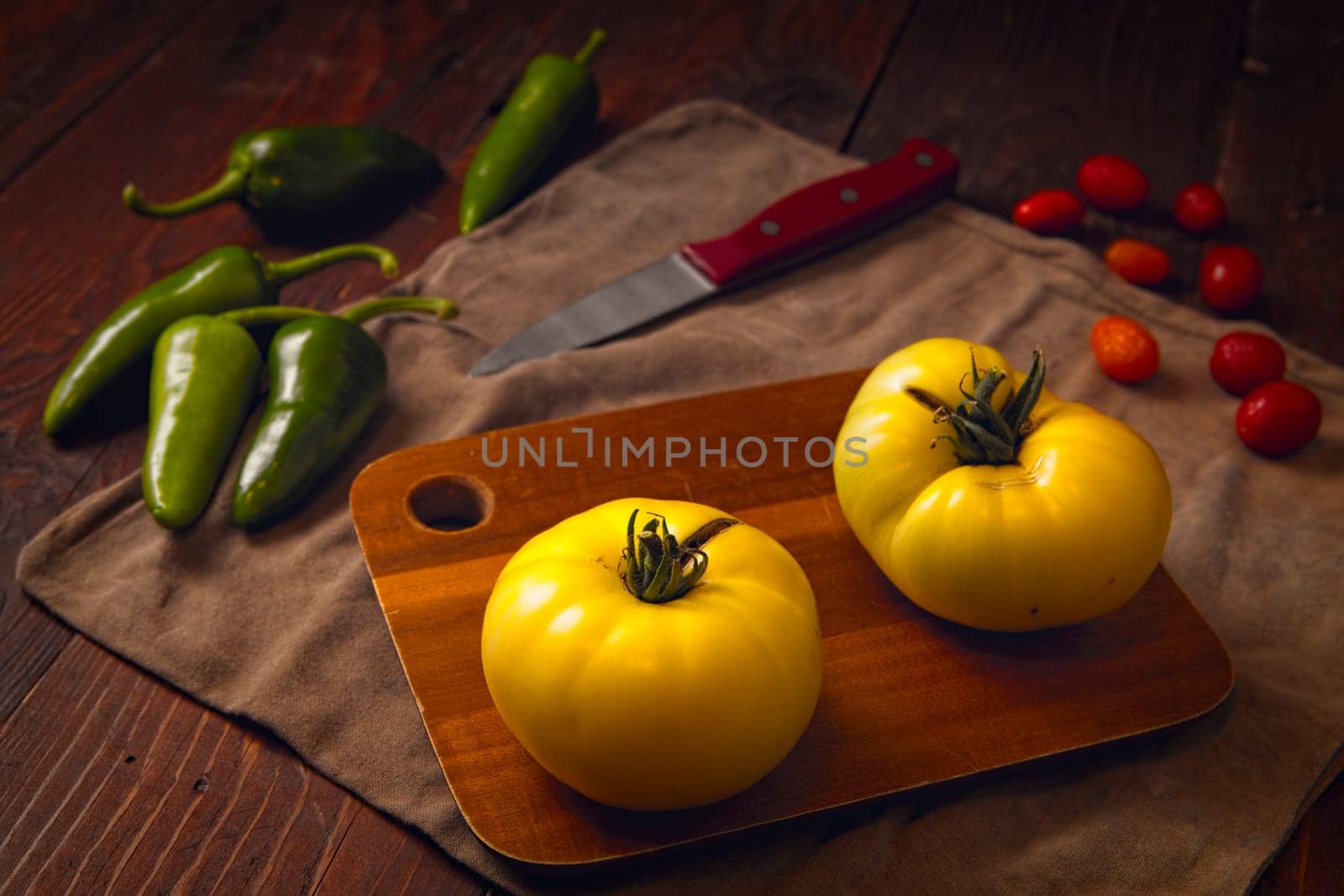 Two ripe yellow tomatoes on a cutting board, spicy green peppers, and some cherry tomatoes.