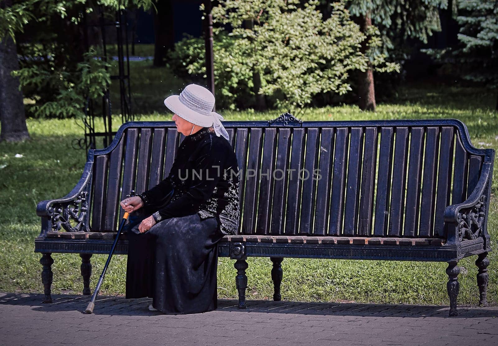 An elderly woman wearing a black dress and white hat sits on a park bench, surrounded by lush trees. The serene expression on her face suggests that she is enjoying the peace and quiet of the park.