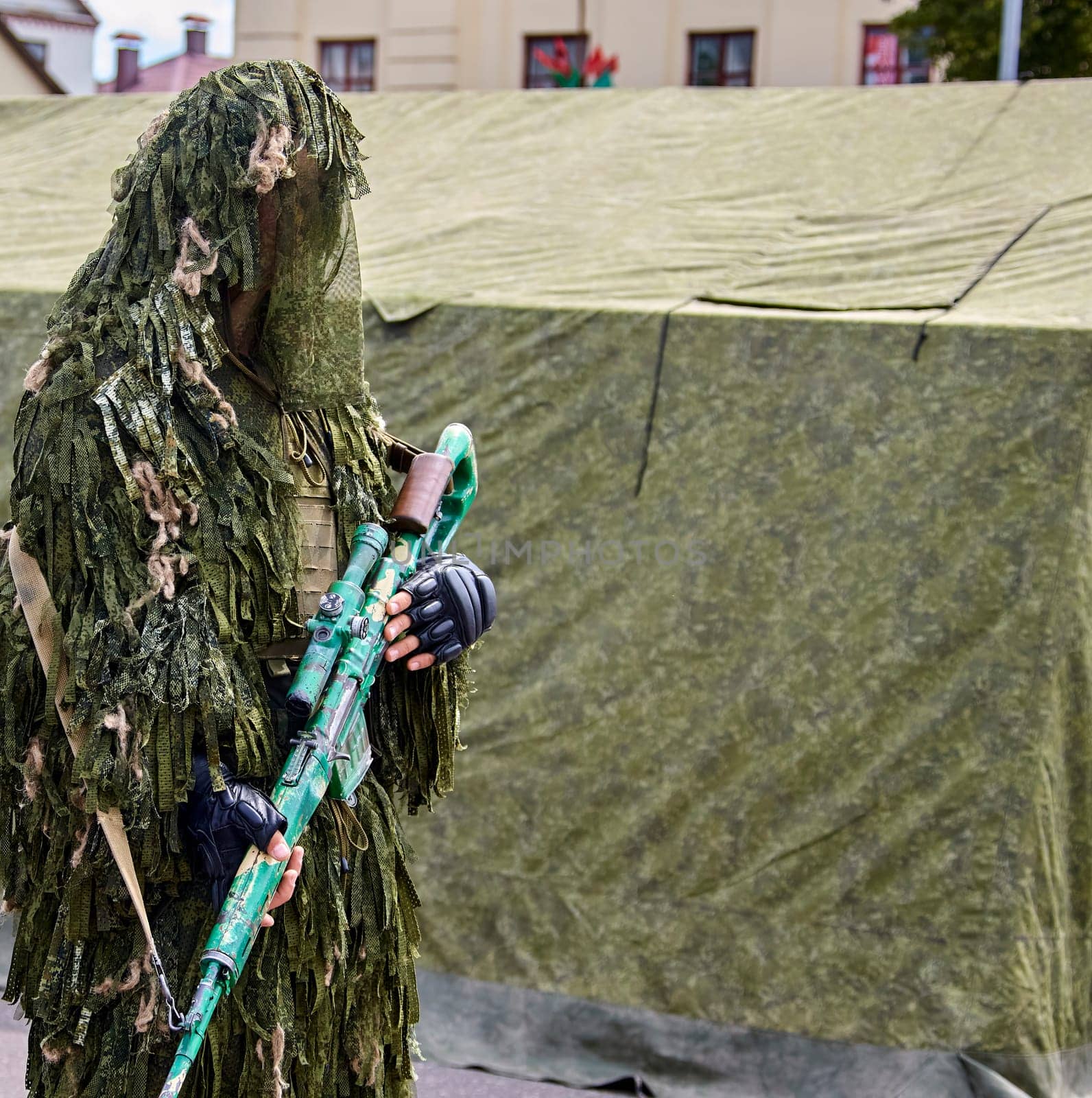 A soldier dressed in camouflage holds a rifle, standing near a green tent.