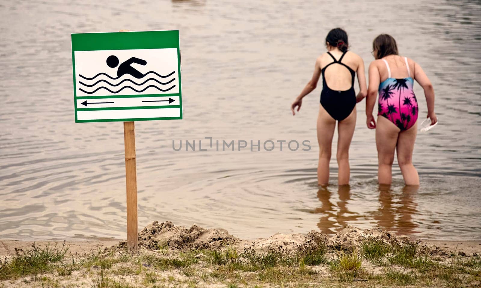 On a bright and sunny afternoon, two young girls stand barefoot in the gentle, shimmering waters of a lake. They are dressed in colorful swimsuits, playfully exploring the shallows while a green sign marks the swimming area nearby.