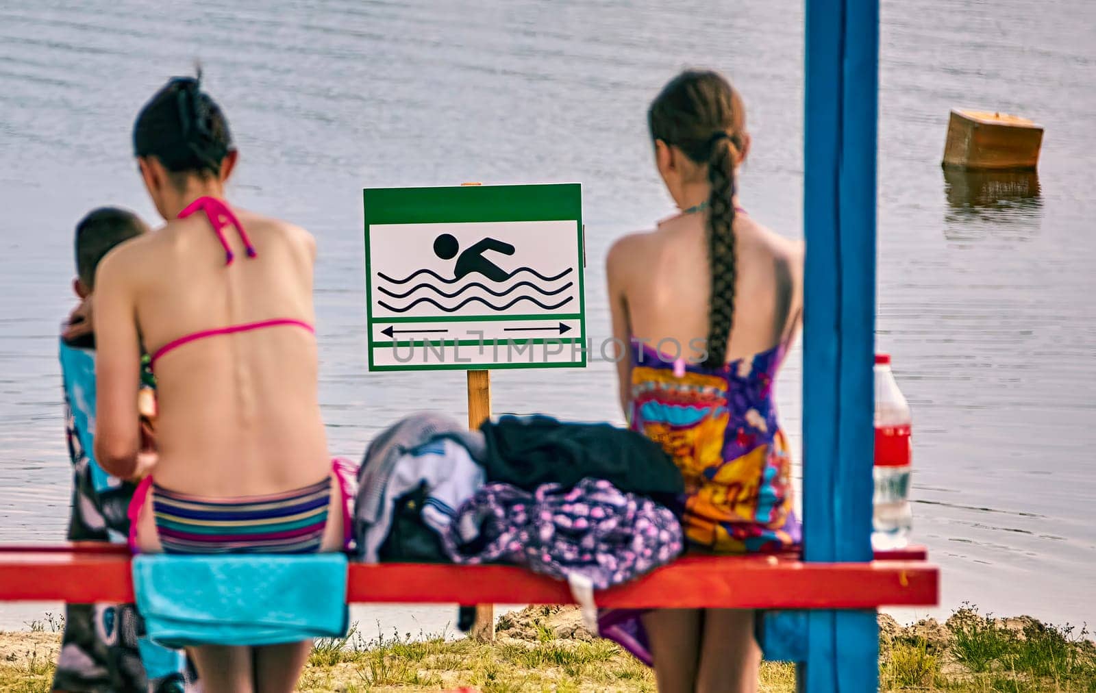 Two individuals sit on a vibrant red bench by a tranquil lake, enjoying the warm weather. They are engaged in conversation, surrounded by colorful towels and swim gear.