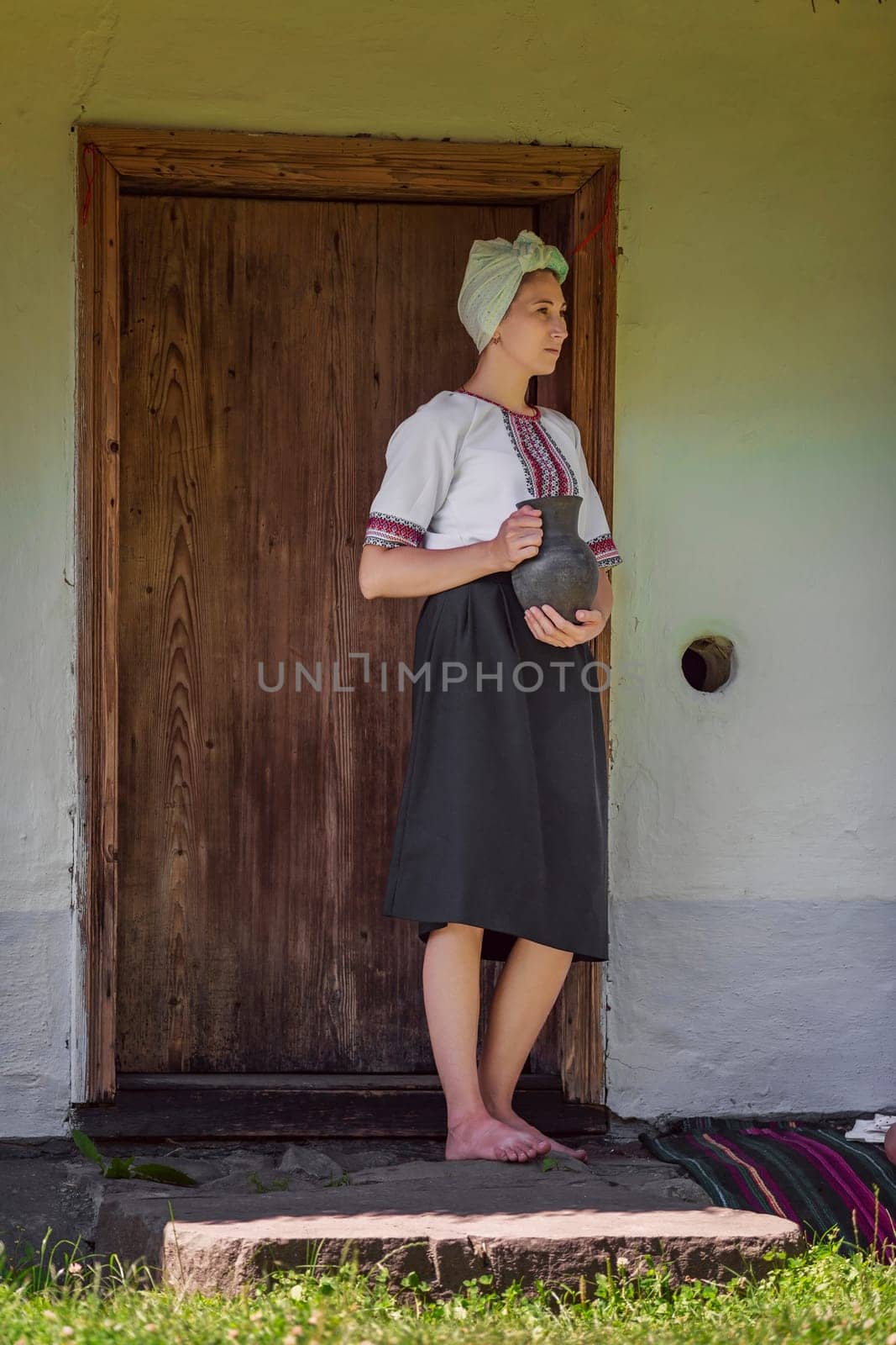 A traditional woman dressed in a beautiful folk costume stands barefoot beside a rustic wooden door