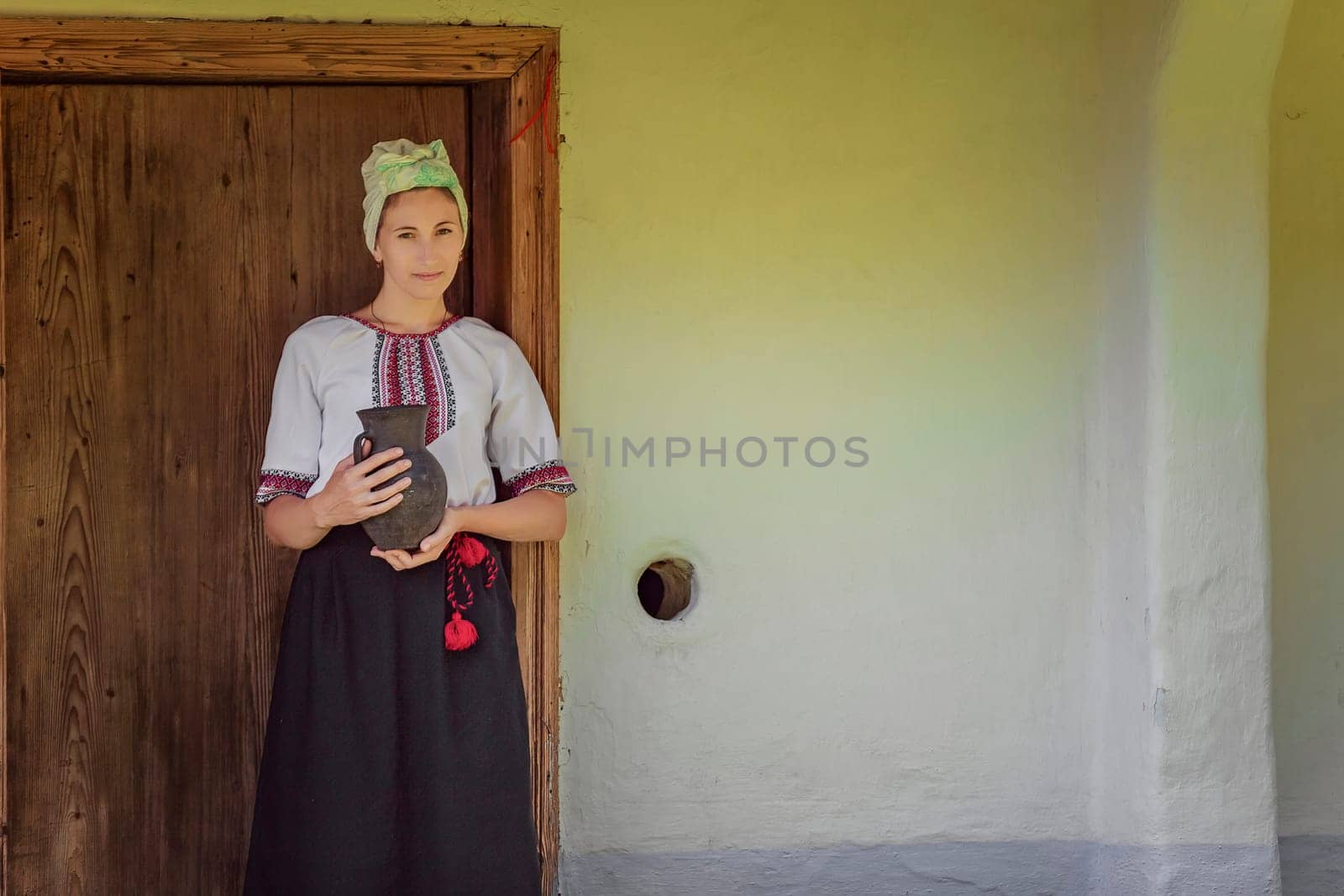 A woman dressed in beautiful traditional attire poses proudly with a decorative pot by an old wooden door