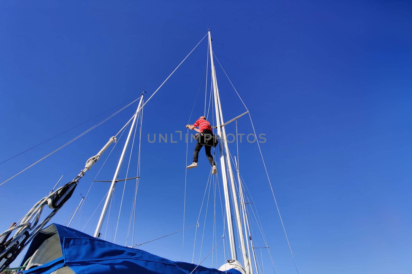 Man working up the mast of a sailing yacht, climbing with rope and bosun's chair on a sunny day with blue sky, repairing, fixing the mast shrouds