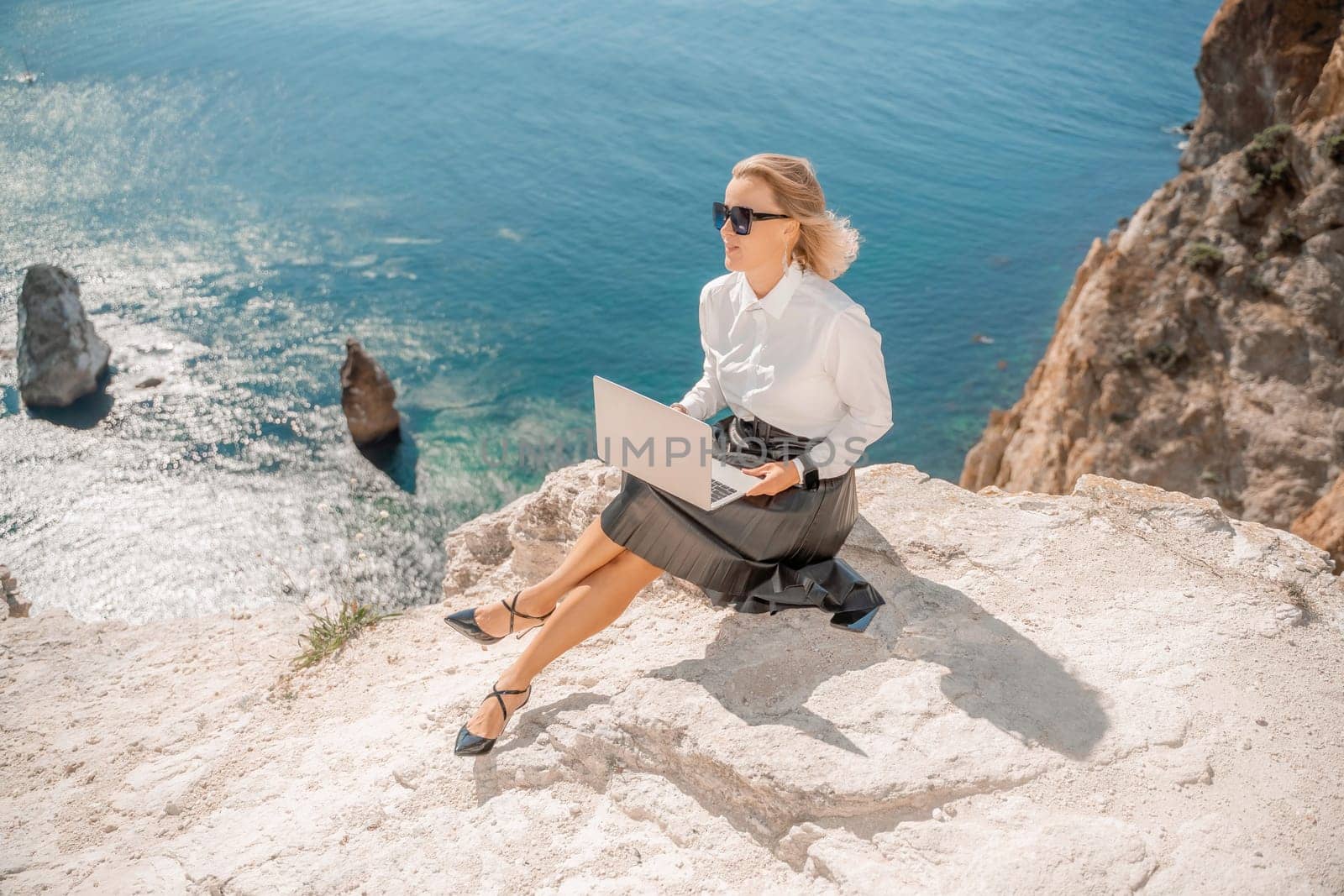 Business woman on nature in white shirt and black skirt. She works with an iPad in the open air with a beautiful view of the sea. The concept of remote work. by Matiunina