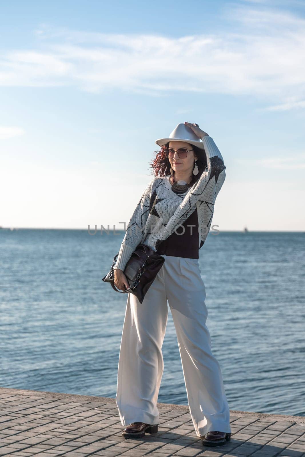 Stylish seashore woman. Fashionable woman in a white hat, white trousers and a light sweater with a black pattern on the background of the sea. by Matiunina