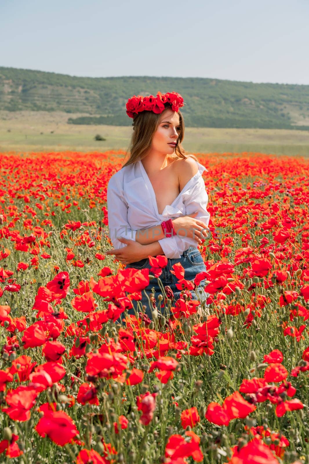 Happy woman in a poppy field in a white shirt and denim skirt with a wreath of poppies on her head posing and enjoying the poppy field. by Matiunina