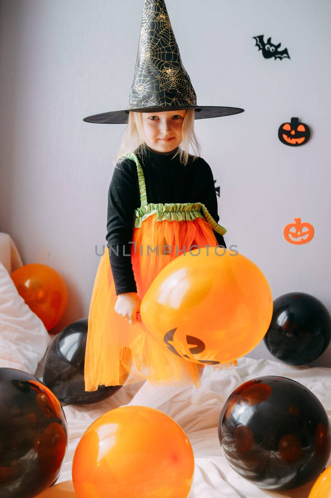 Children's Halloween - a girl in a witch hat and a carnival costume with airy orange and black balloons at home. Ready to celebrate Halloween
