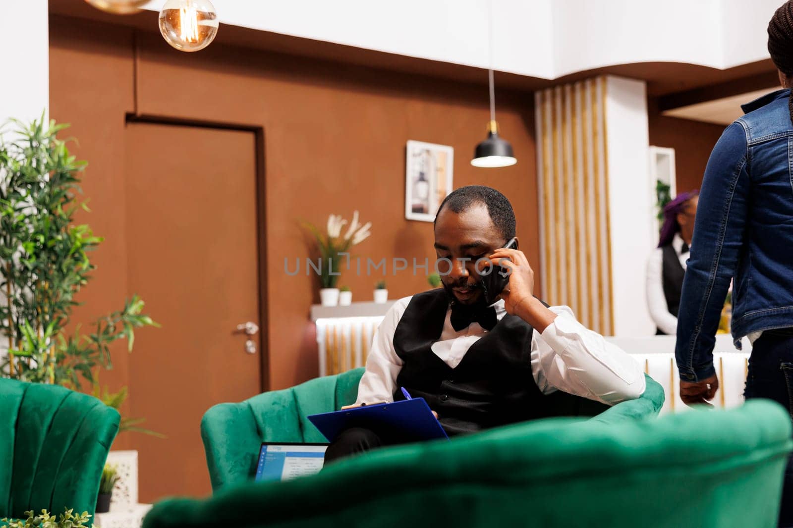 African American man hotel bellboy sitting in lobby talking on phone and taking notes. Concierge calling cab contacting transportation service, assisting guests in arranging for transportation