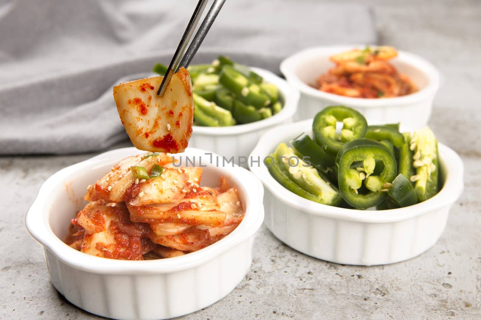 A food photo of a pair of chopsticks holding a piece of kimchi above bowls of kimchi and sliced peppers.
