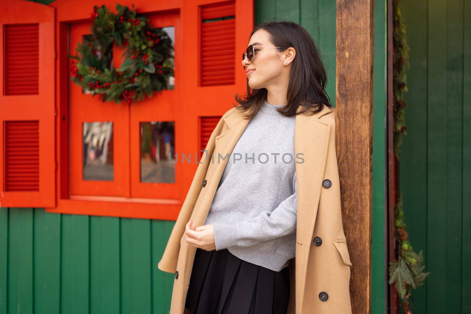 People, season and Christmas concept portrait of happy smiling middle age woman outdoors dressed stylish cream trench coat with Christmas festive decoration on background. Looking away, copy space