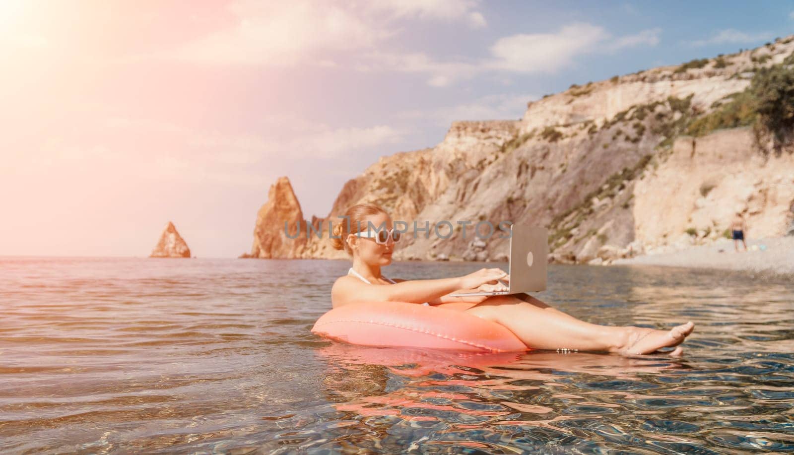Woman freelancer works on laptop swimming in sea on pink inflatable ring. Pretty lady typing on computer while floating in the sea on inflatable donut at sunset. Freelance, remote work on vacation