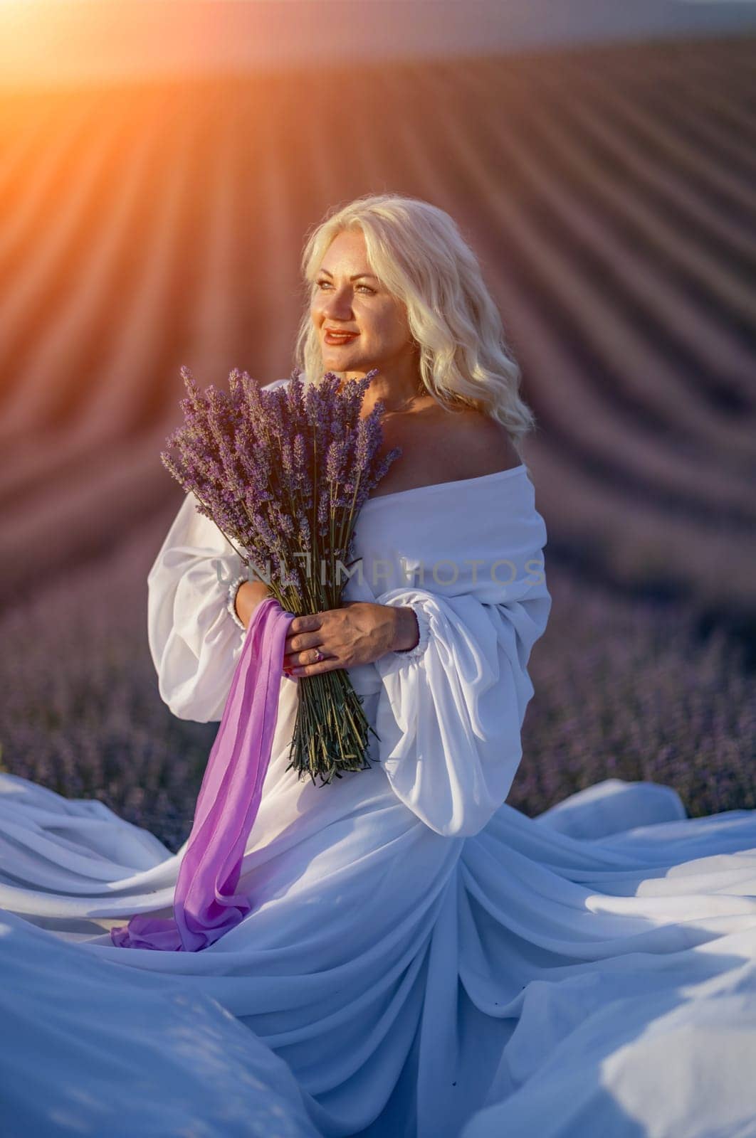 Blonde woman poses in lavender field at sunset. Happy woman in white dress holds lavender bouquet. Aromatherapy concept, lavender oil, photo session in lavender.