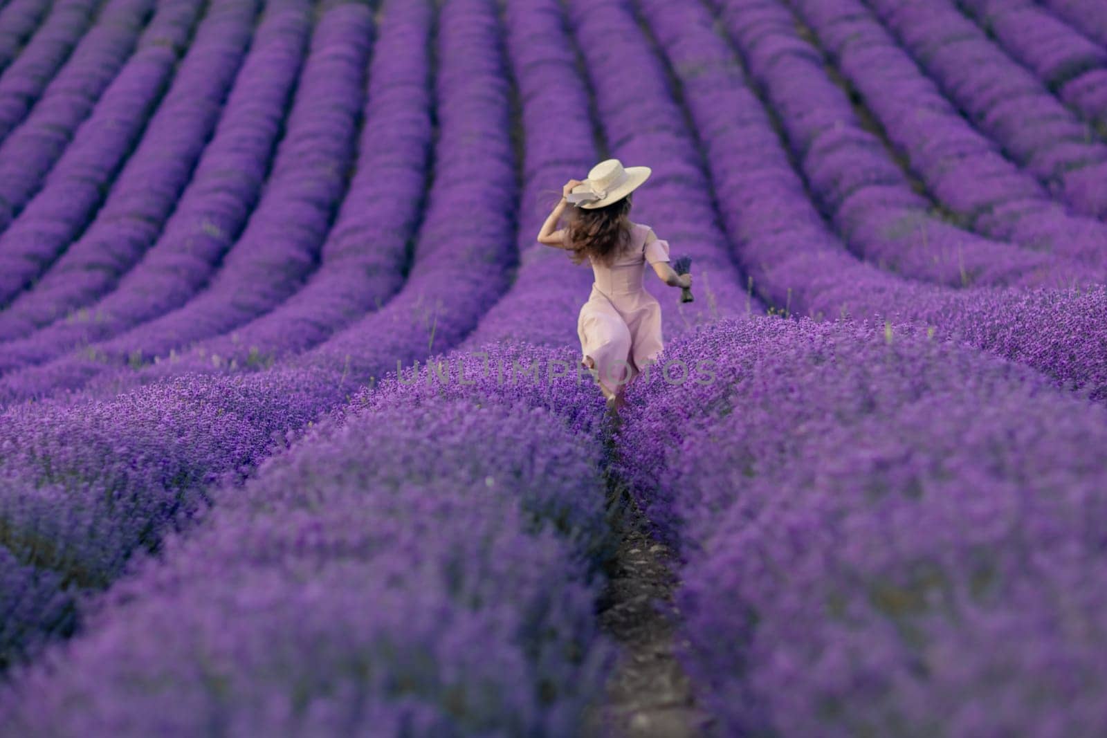 A woman is walking through a field of purple flowers. The field is vast and the flowers are in full bloom. The woman is wearing a hat and a pink dress