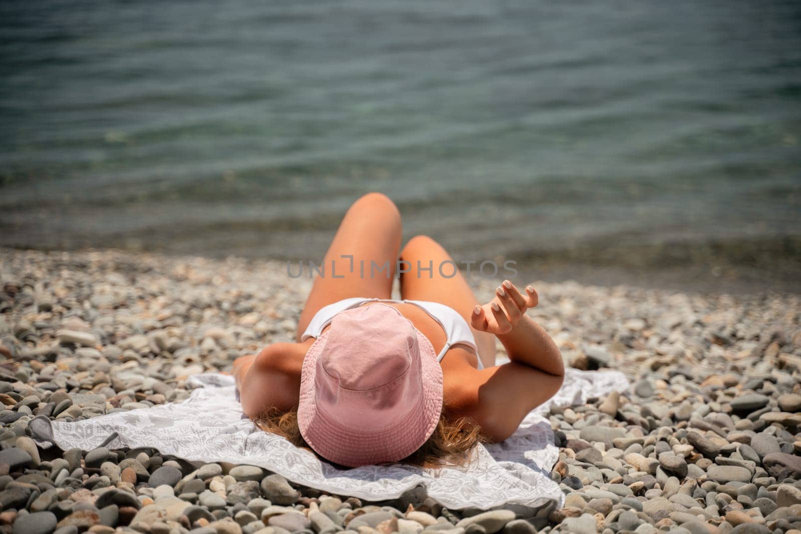 A woman is laying on the beach with a pink hat on her head. She is wearing a white bikini and is laying on a towel. The beach is rocky and the water is calm. Scene is relaxed and peaceful