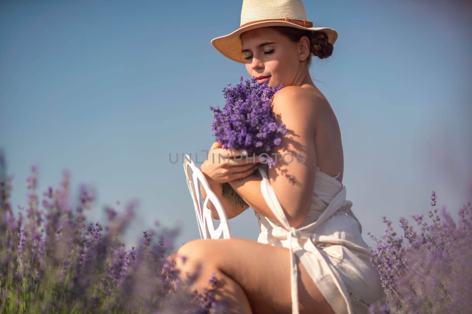 A woman is sitting in a field of lavender flowers and wearing a straw hat. She is smiling and holding a bouquet of flowers. Scene is peaceful and serene, as the woman is surrounded by the beauty of nature.