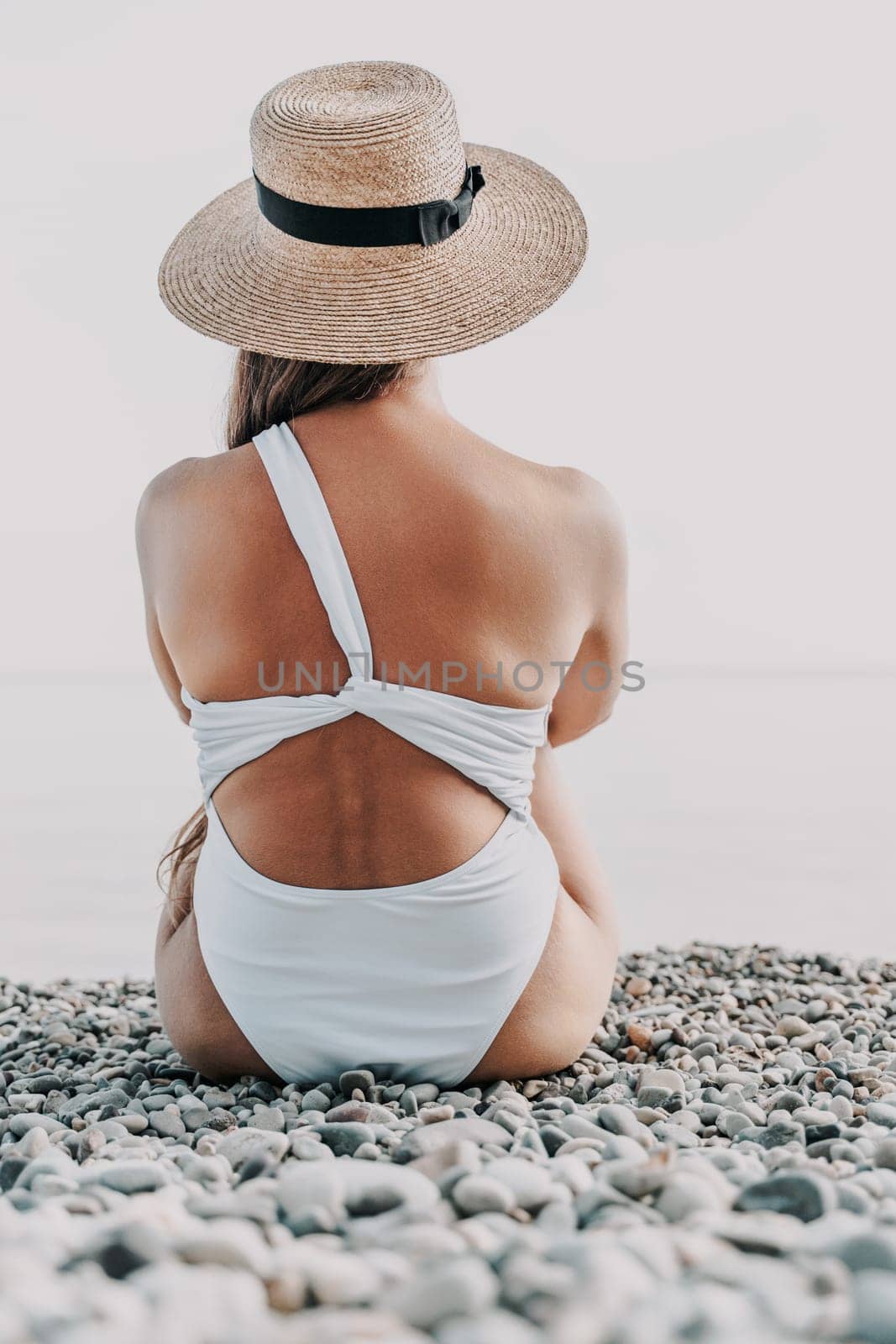 A woman is sitting on a beach wearing a straw hat and a white bikini. The beach is rocky and the sky is blue
