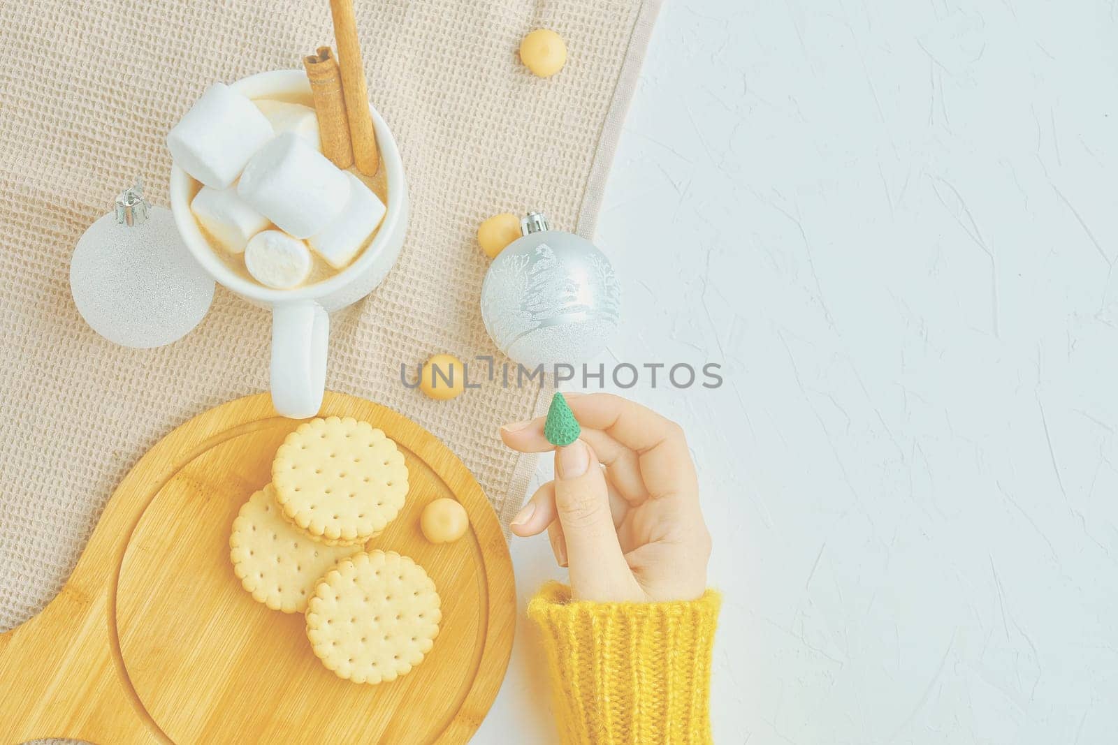 Woman in a yellow sweater drinks hot cocoa with marshmallow and cinnamon rolls in a white mug and cookies and a figure of a Christmas tree