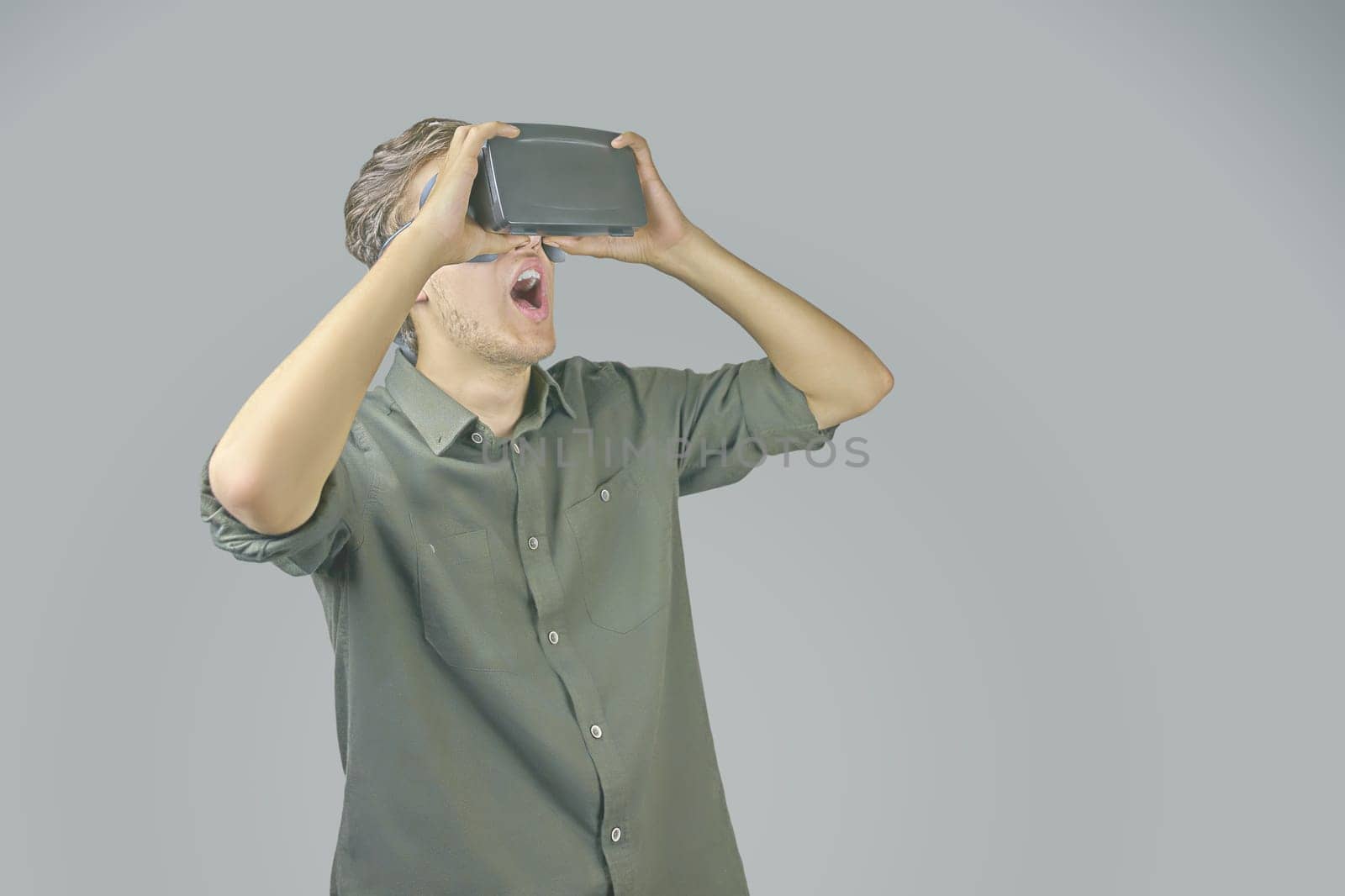 Young man in shirt enjoys and amazed by virtual reality glasses in front of light grey background