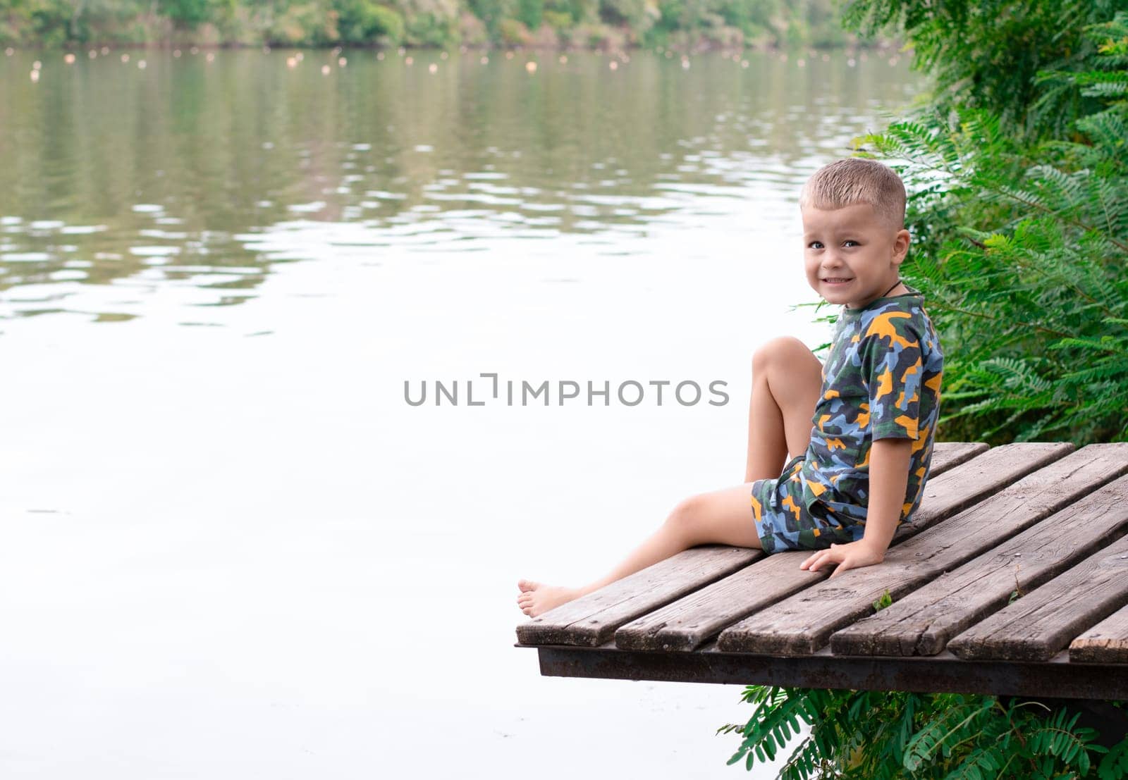 Fishing concept. A small handsome boy, with emotional facial expressions, 4 years old, sits on a wooden bridge by the river, against the backdrop of green plants and breathes fresh air. Relax. Place for text. Copy space. Vacations, Village.