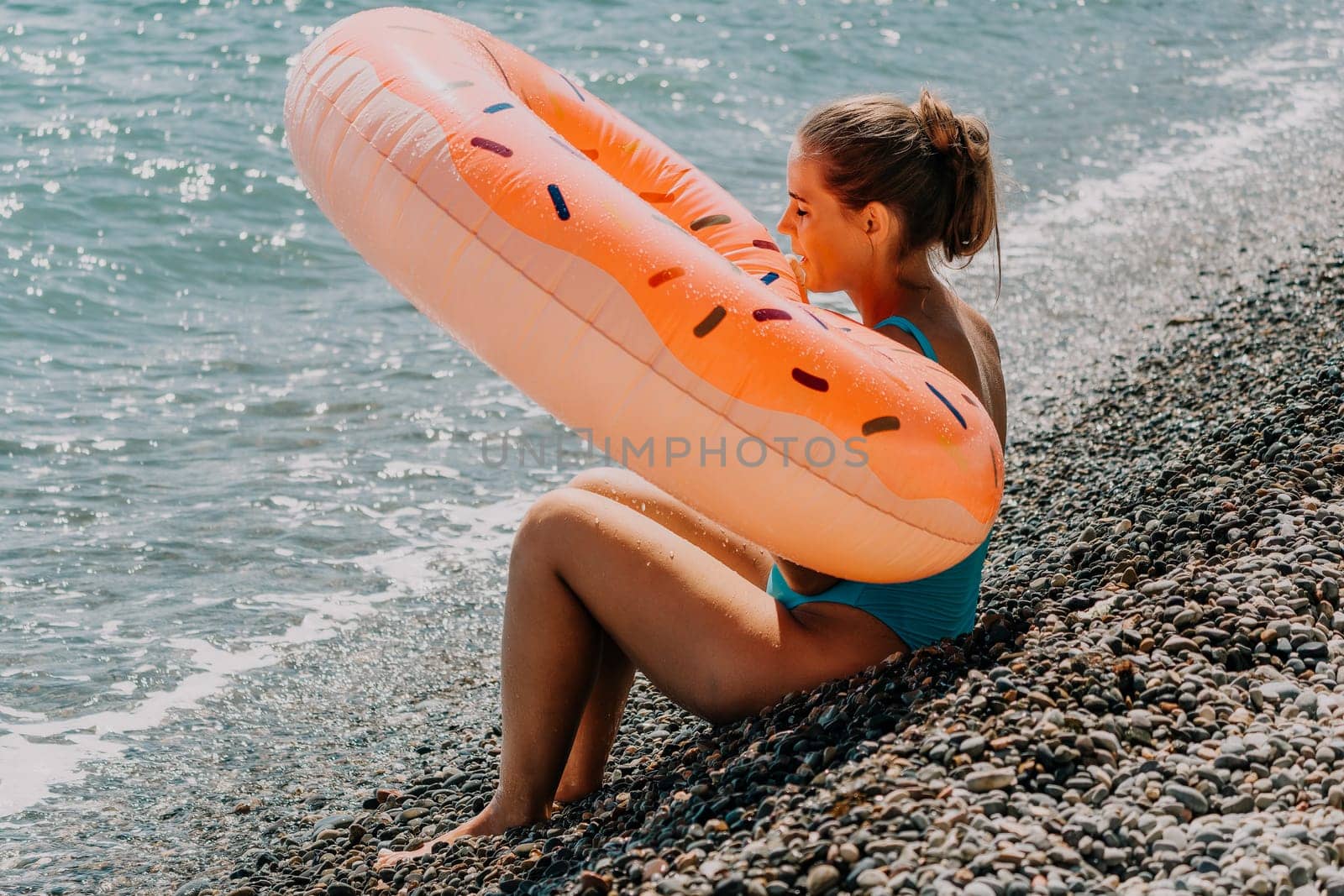A young woman in a swimsuit sits on a pebble beach with a donut-shaped float behind her.