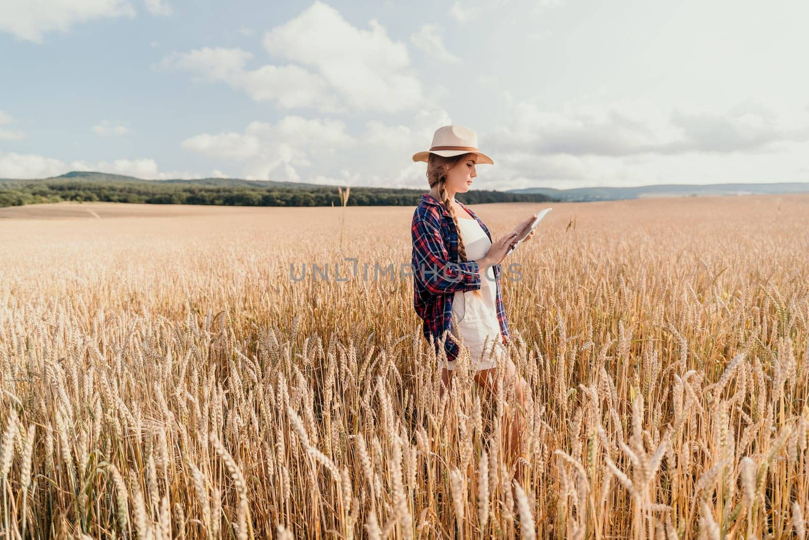 A young woman in a hat and plaid shirt stands in a wheat field and uses a tablet.