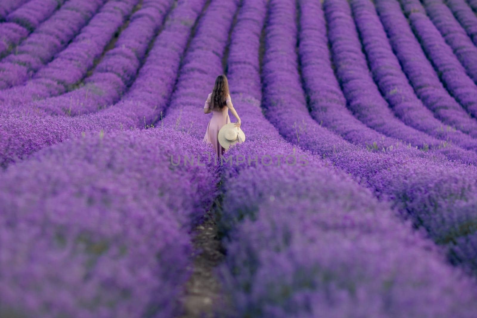 A woman is walking through a field of purple flowers. She is wearing a white hat and a pink dress. The scene is peaceful and serene