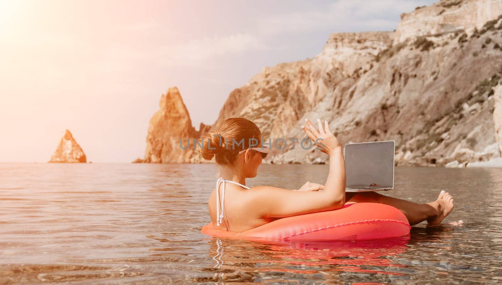 Woman freelancer works on laptop swimming in sea on pink inflatable ring. Pretty lady typing on computer while floating in the sea on inflatable donut at sunset. Freelance, remote work on vacation