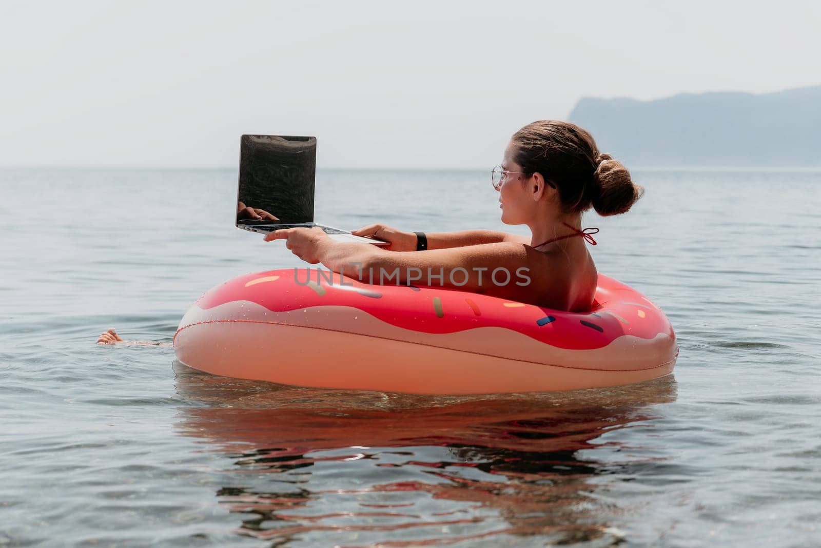 Woman freelancer works on laptop swimming in sea on pink inflatable ring. Pretty lady typing on computer while floating in the sea on inflatable donut at sunset. Freelance, remote work on vacation