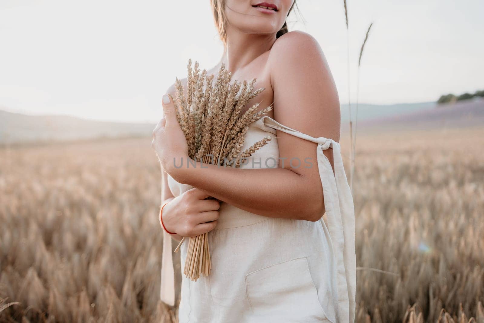 A woman is holding a bunch of wheat in her arms. The wheat is dry and brown, and the woman is wearing a white dress. The scene is set in a field, and the woman is posing for a photo