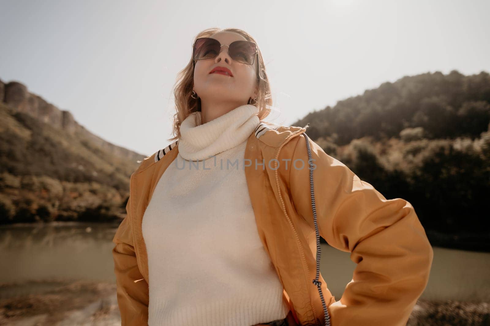 A young woman in a yellow jacket and sunglasses stands by a lake, with hills in the background and a blue sky overhead.