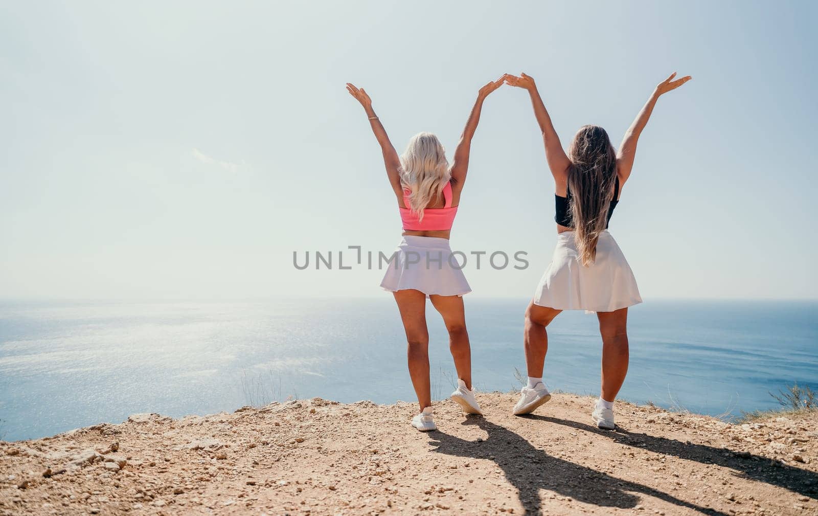 Two women stand on a cliffside with their backs to the camera, their arms raised in a triumphant pose. The ocean stretches out before them under a clear blue sky.