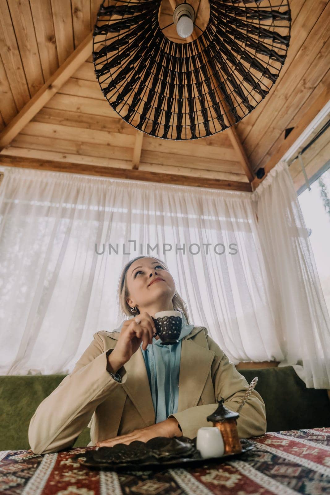 A woman dressed in a beige jacket enjoys Turkish coffee in a cozy cafe, gazing up at the intricate ceiling lamp.