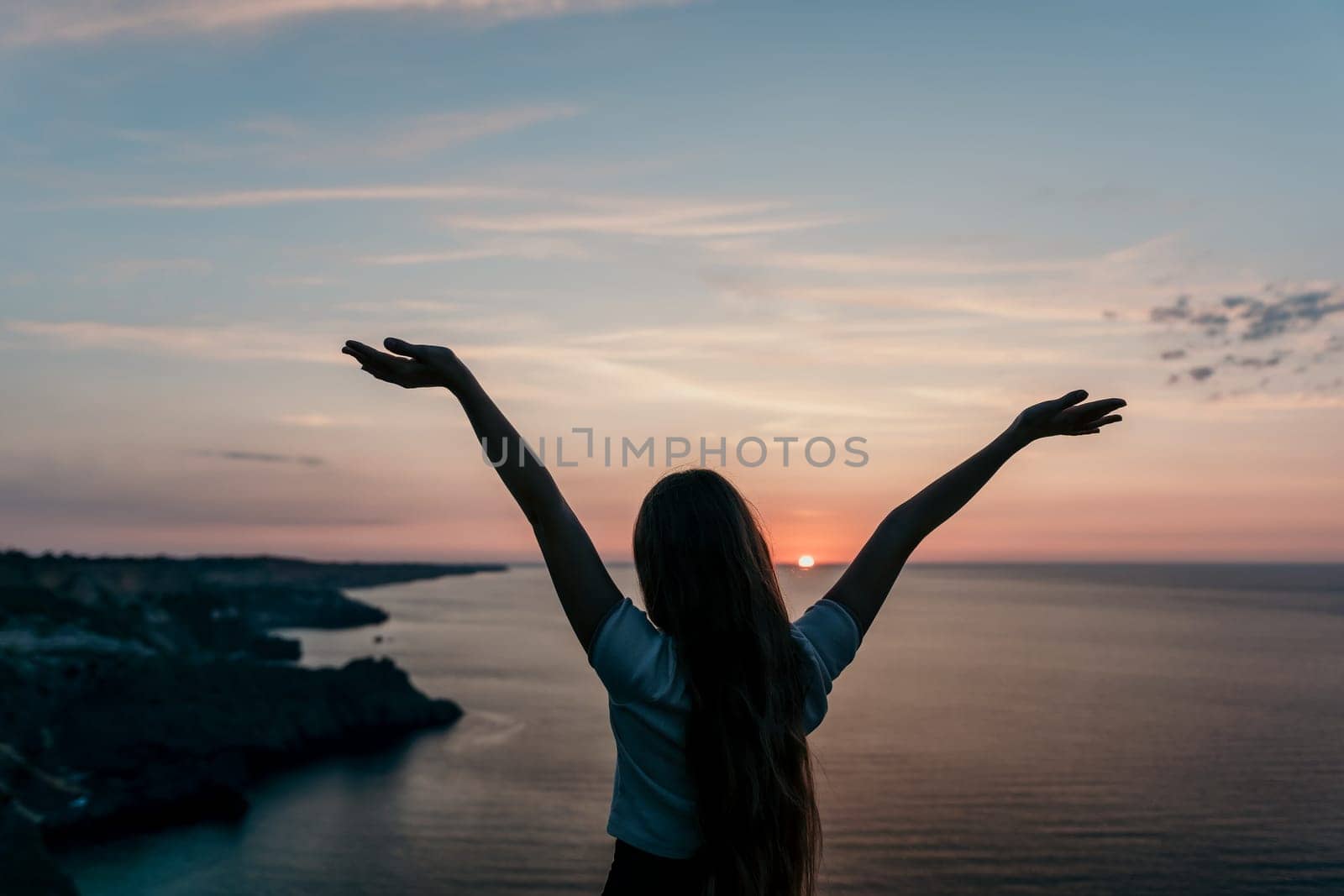 A woman stands with open arms, her silhouette outlined against a vibrant sunset over the ocean.