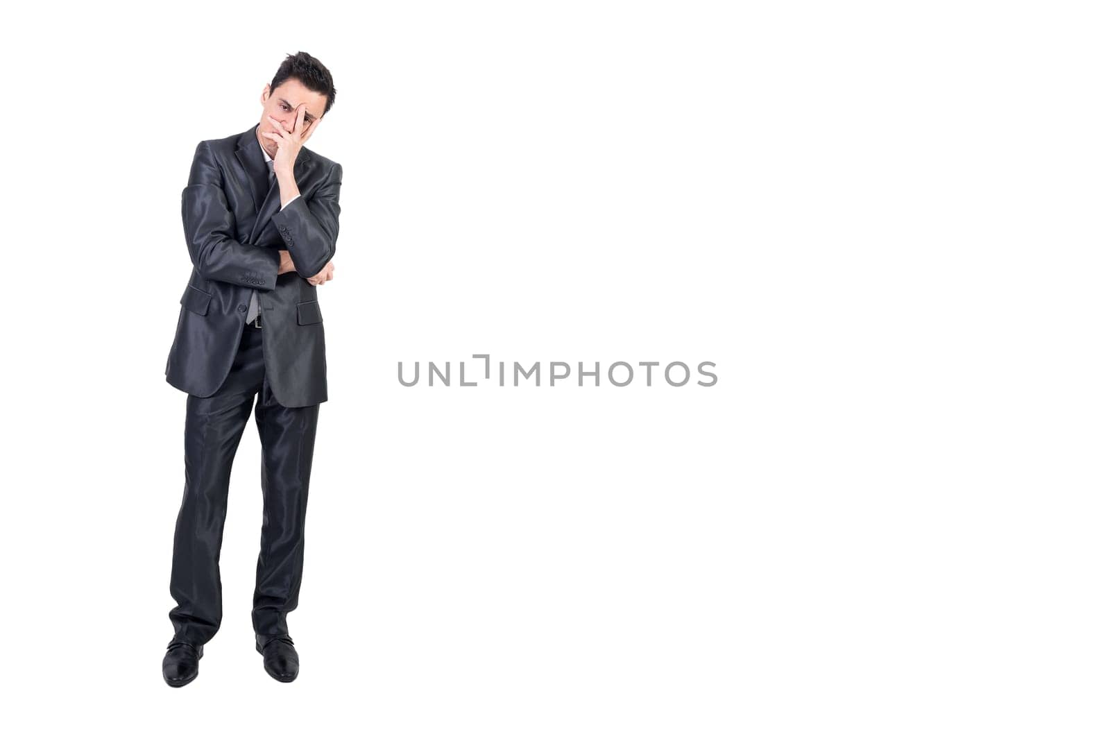 Full length of bored young male entrepreneur with dark hair in formal suit covering face with hand and looking at camera against white background