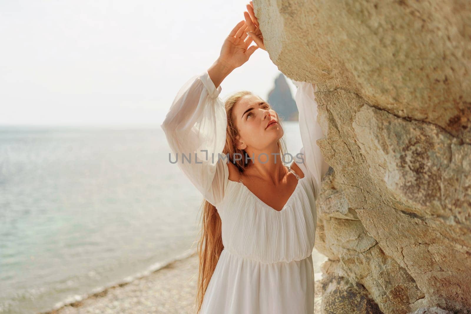 woman white dress standing on a beach, looking out at the ocean. She is lost in thought, with her hand on her hip. Concept of calm and introspection