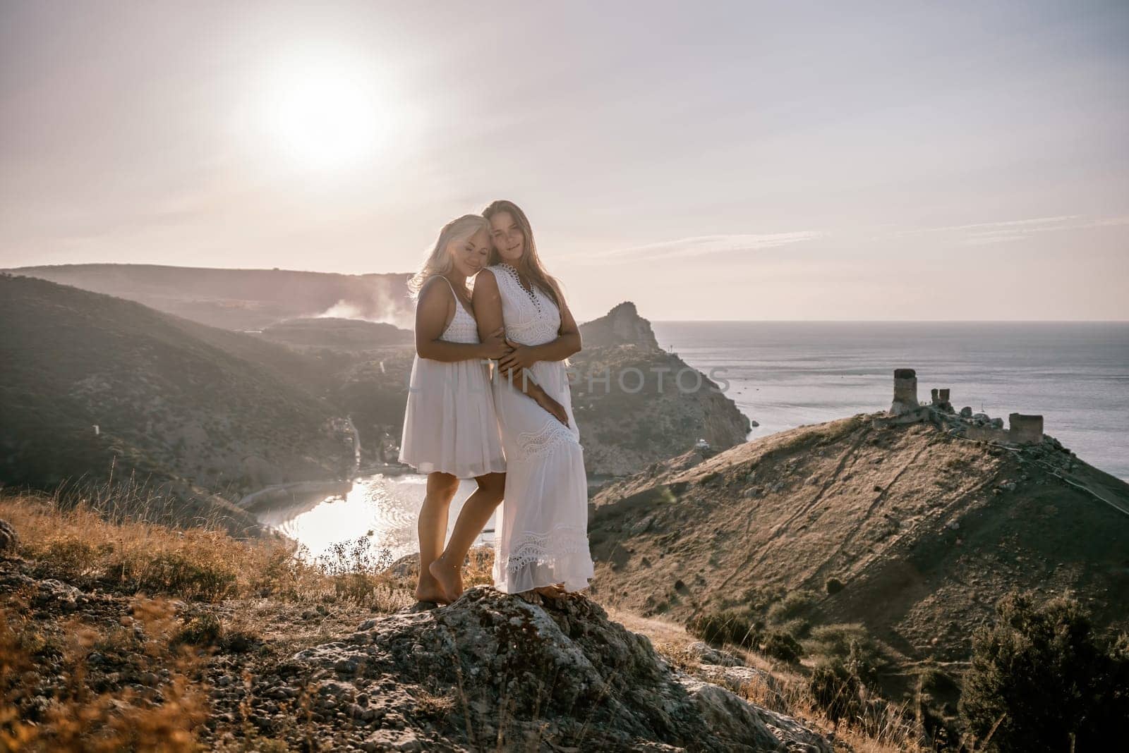 Two young girls are standing on a hillside, one of them wearing a white dress. The sun is shining brightly, creating a warm and inviting atmosphere. The girls seem to be enjoying their time together