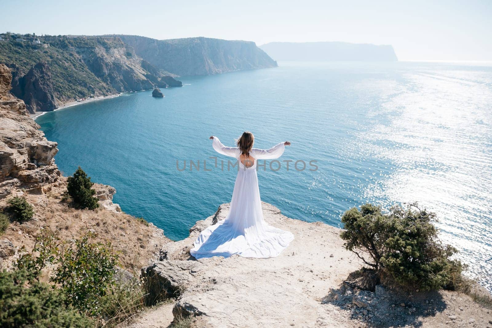 A woman stands on a cliff overlooking the ocean in a white dress. She is looking out at the water and she is in a state of peace and serenity. Concept of calm and tranquility