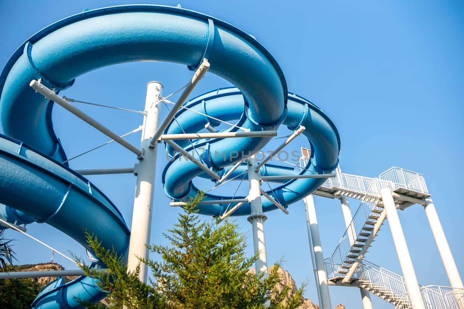 A blue waterslide twists and turns against a clear blue sky. The slide is made of metal and has a white frame. A small green tree is visible in the lower left corner.