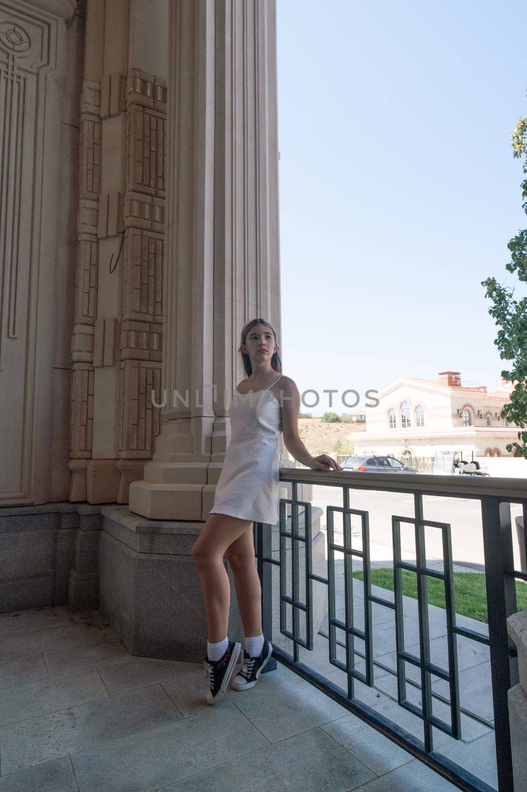 A young woman in a white dress leans on a railing outside a building with columns. She looks towards the right side of the frame.