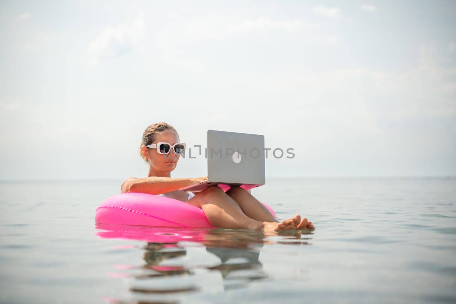 A woman is sitting on a pink inflatable raft in the ocean, using a laptop. Concept of relaxation and leisure, as the woman is enjoying her time in the water while working on her laptop